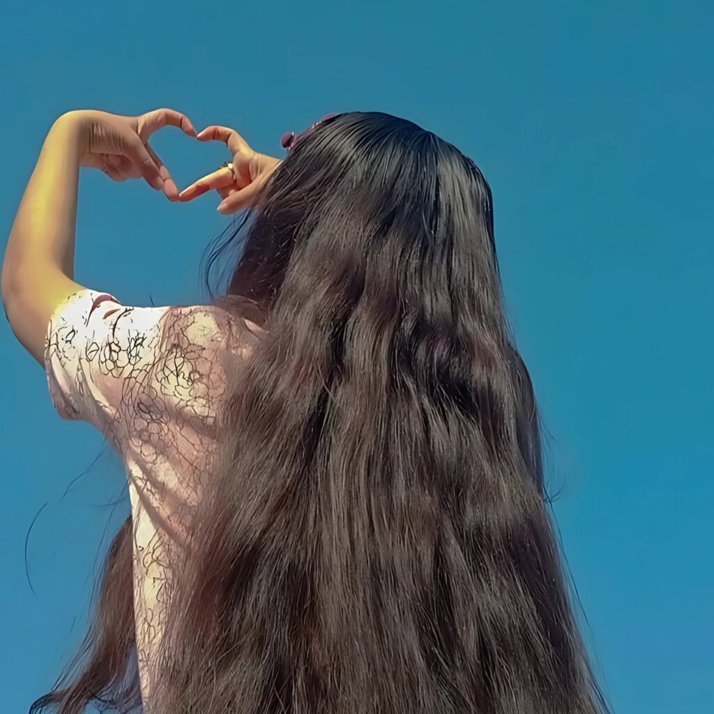 Hidden girl making heart shape with hands under clear blue sky, long wavy hair flowing down her back.
