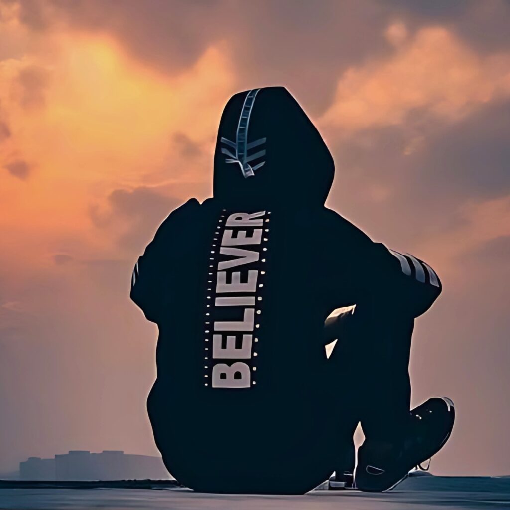 Boy wearing a hoodie with "BELIEVER" written on the back, sitting alone under a dramatic sunset sky — symbolizing strength, belief, and calm attitude.
