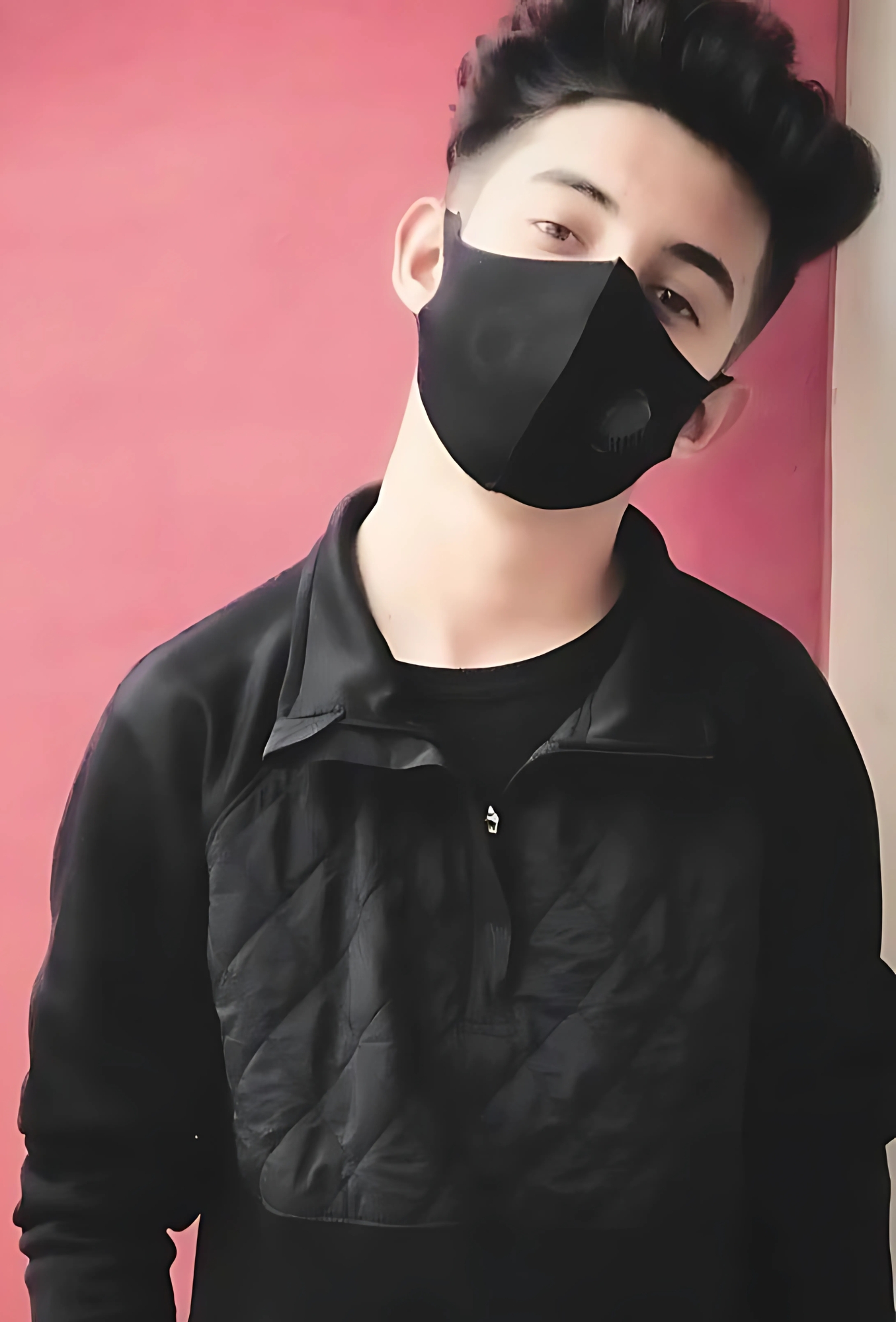 Young boy wearing a black mask and black sweatshirt standing against a pink wall