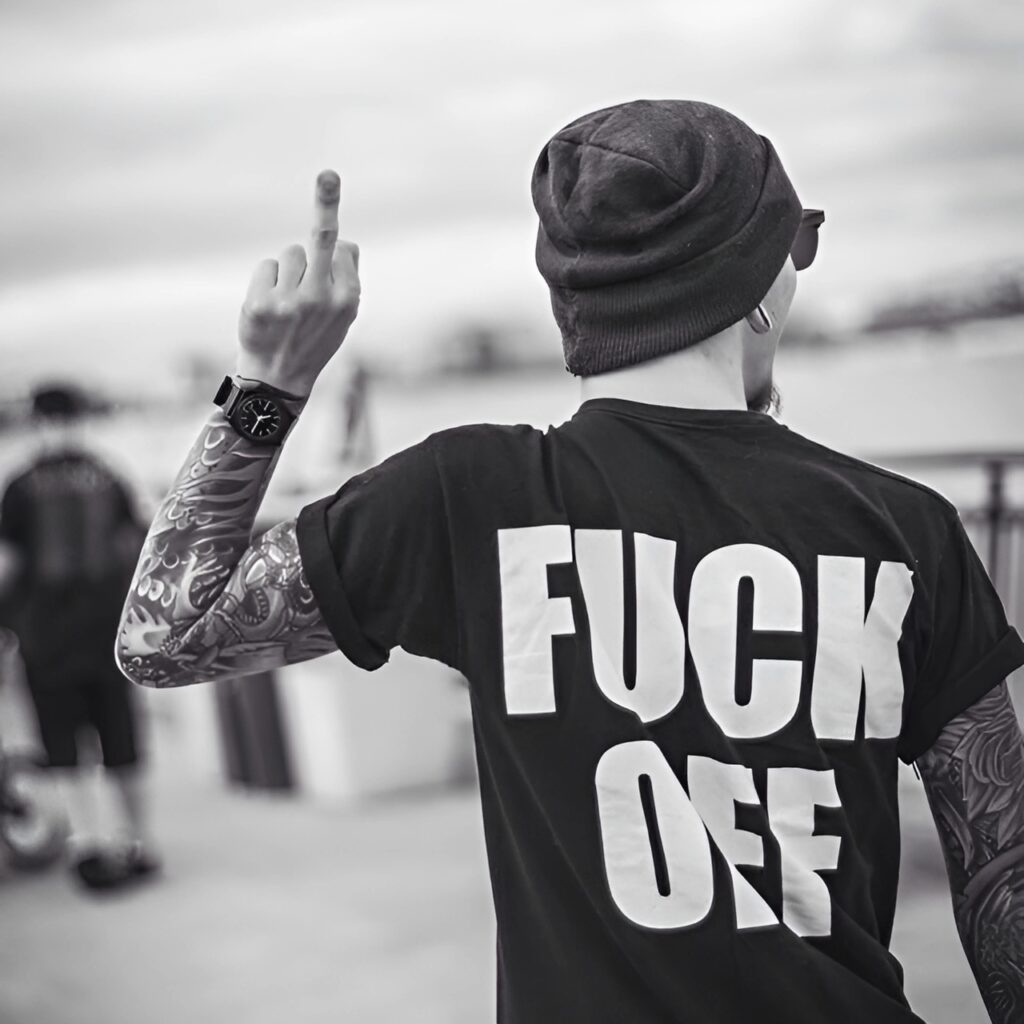 Tattooed boy wearing a bold t-shirt with a rebellious message, showing attitude by flipping the middle finger – perfect for strong statement WhatsApp DPs.