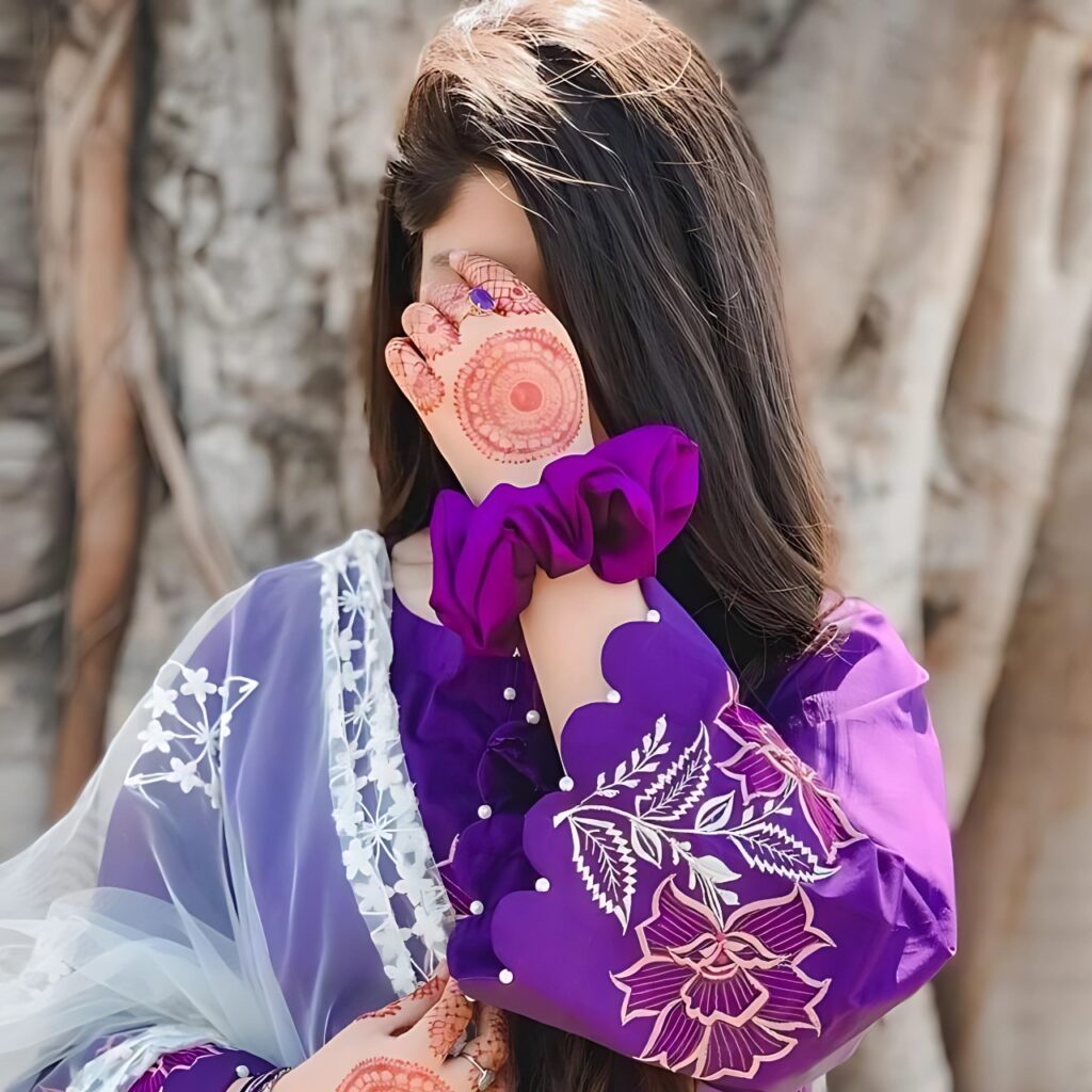Girl hiding face with mehndi-decorated hand, wearing a purple embroidered dress and white net dupatta, standing in front of a tree