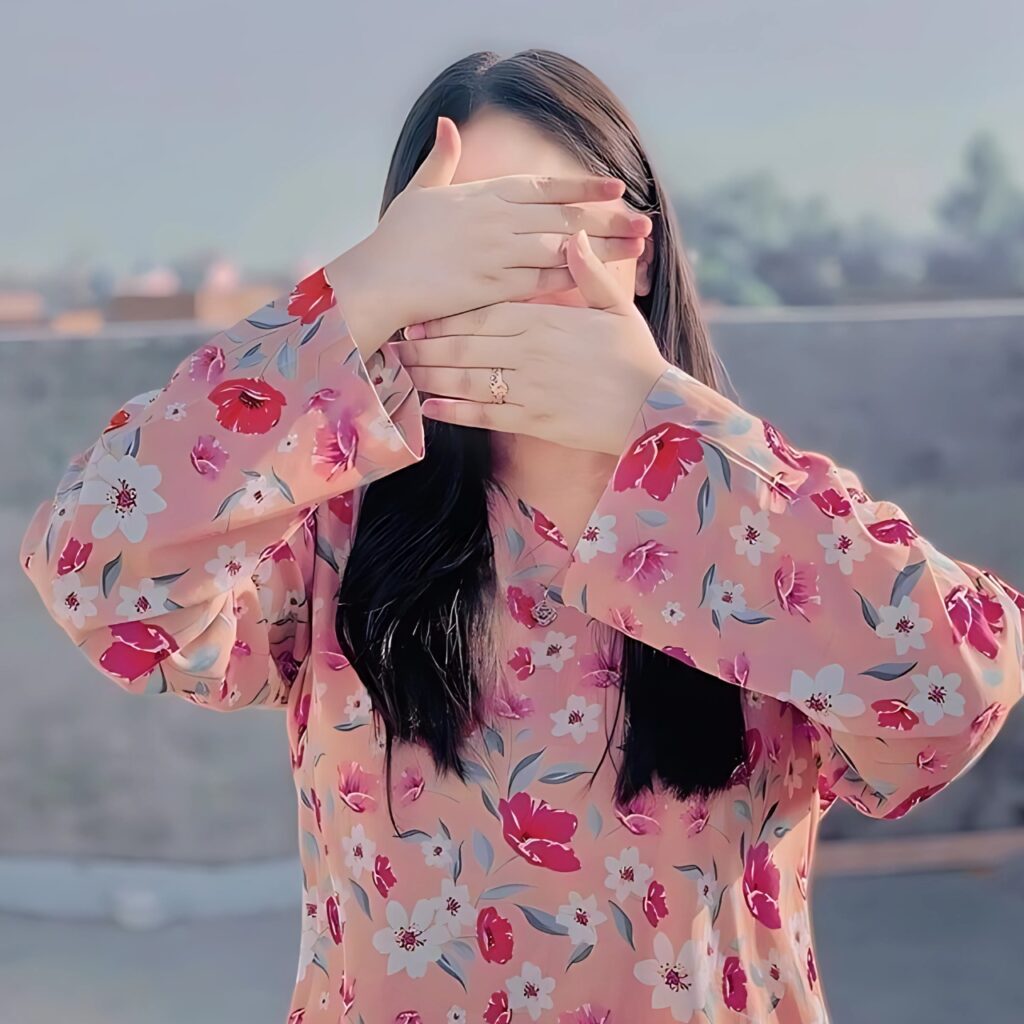 Girl with face hidden using hands, wearing a pink floral dress, standing outdoors under soft daylight