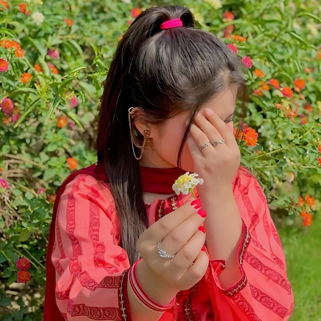 Indian girl hiding face while holding small white flowers, wearing red traditional outfit, sitting in a garden with colorful flower