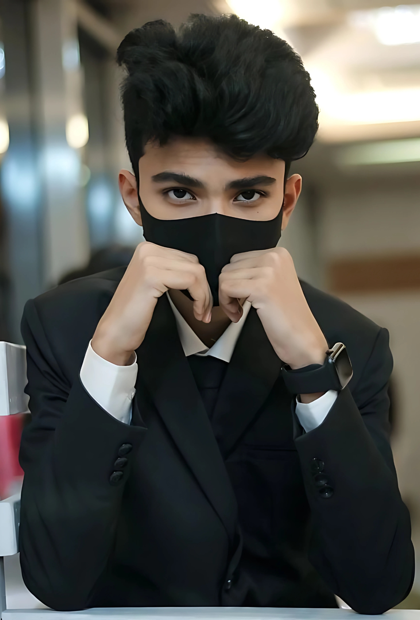 HD photo of a stylish boy in a formal black suit and face mask sitting indoors with intense expression