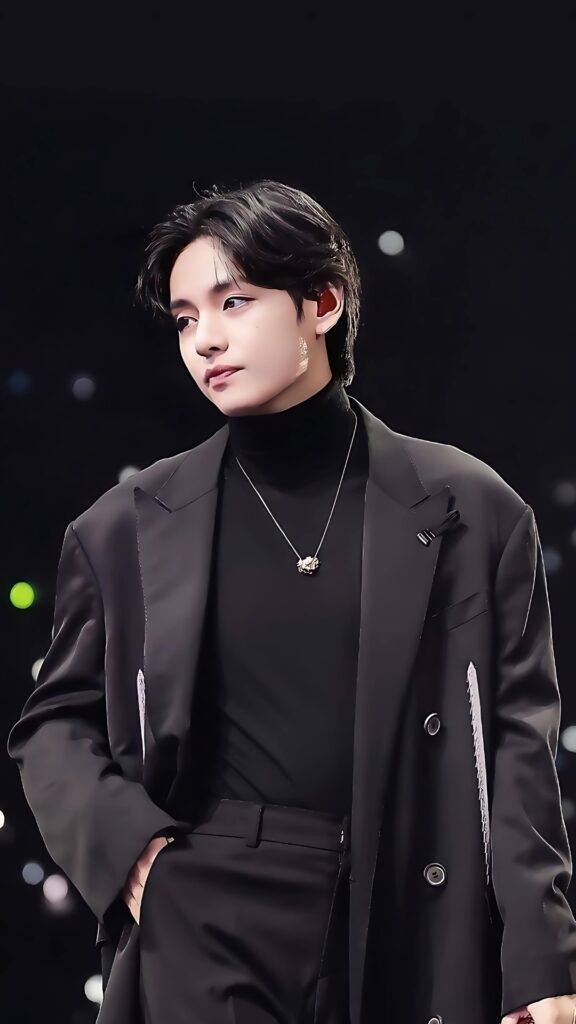 HD wallpaper of BTS member V (Kim Taehyung) in an elegant black suit with a pendant necklace, striking a charismatic pose on stage with a dark bokeh background