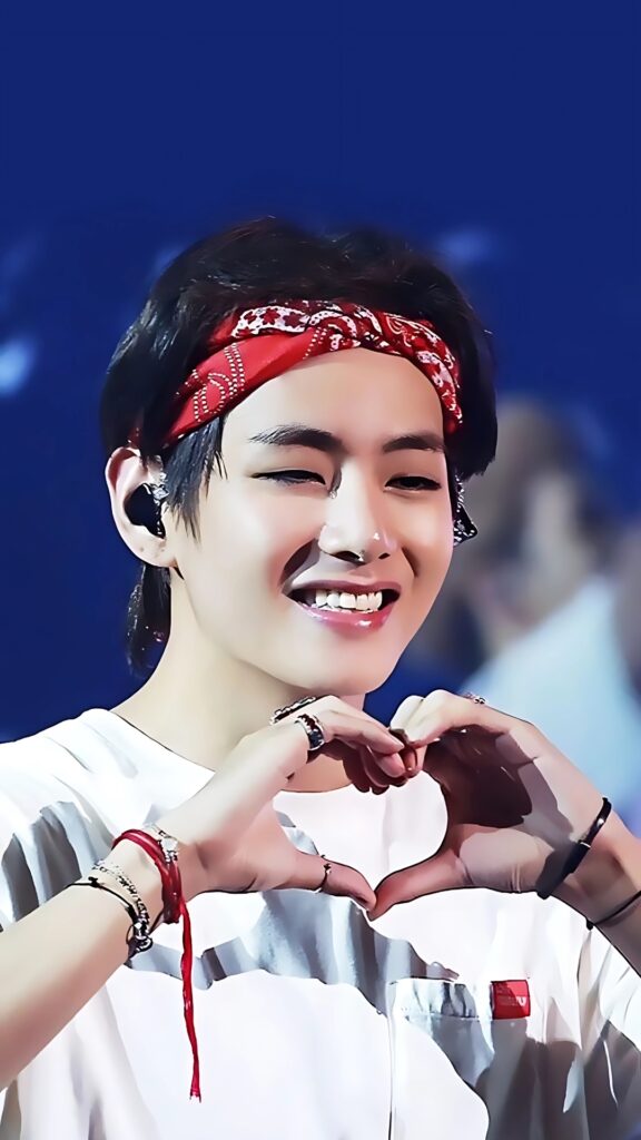 BTS V smiling with a wink and making a heart pose, wearing a red bandana and white shirt on stage