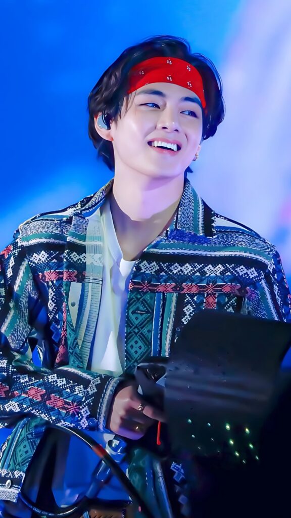 BTS member V smiling brightly on stage, wearing a red bandana and colorful patterned jacket