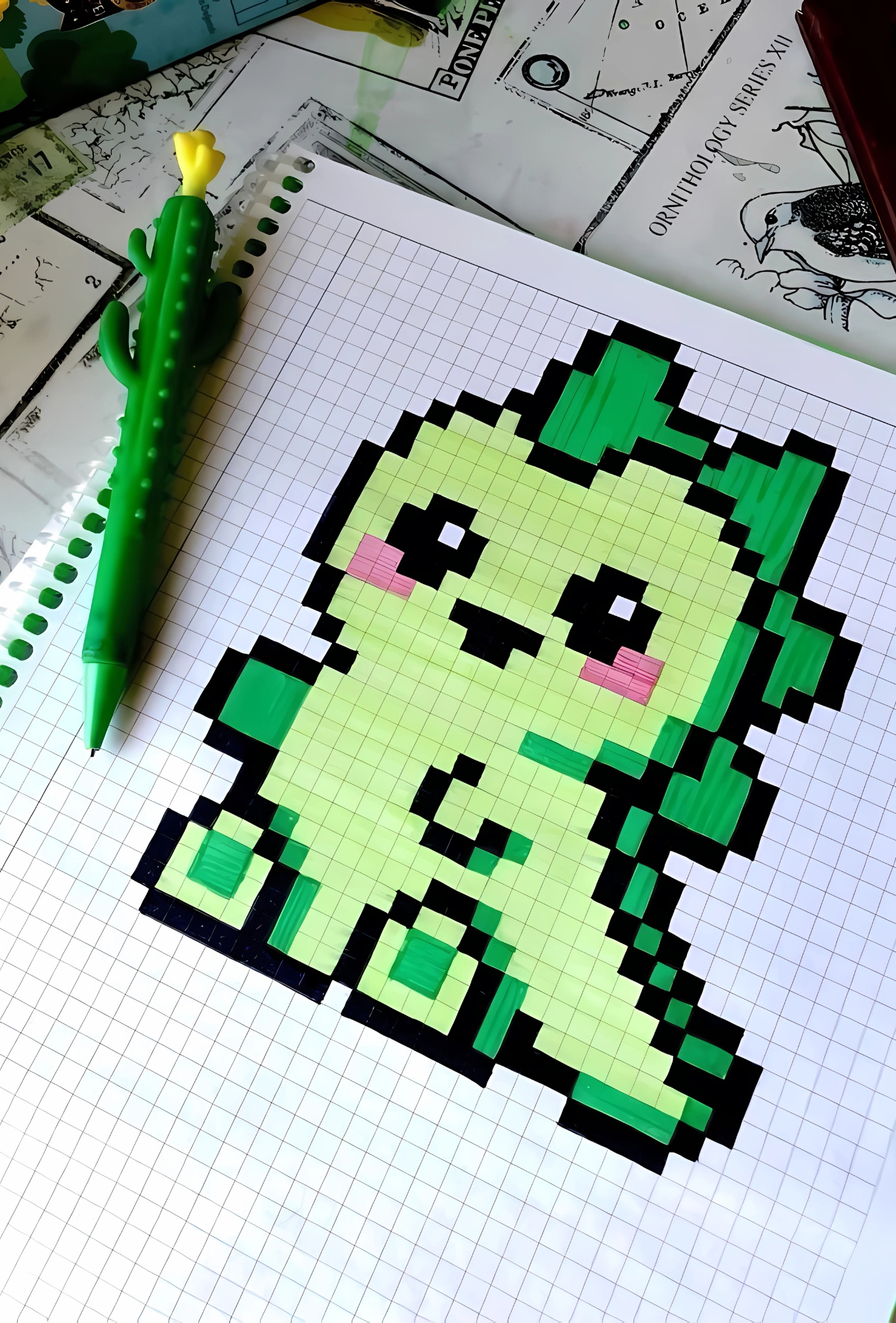 Dibujo pixelado fácil y bonito de personaje tipo planta kawaii hecho con marcadores verdes en cuaderno de cuadros, junto a bolígrafo en forma de cactus.