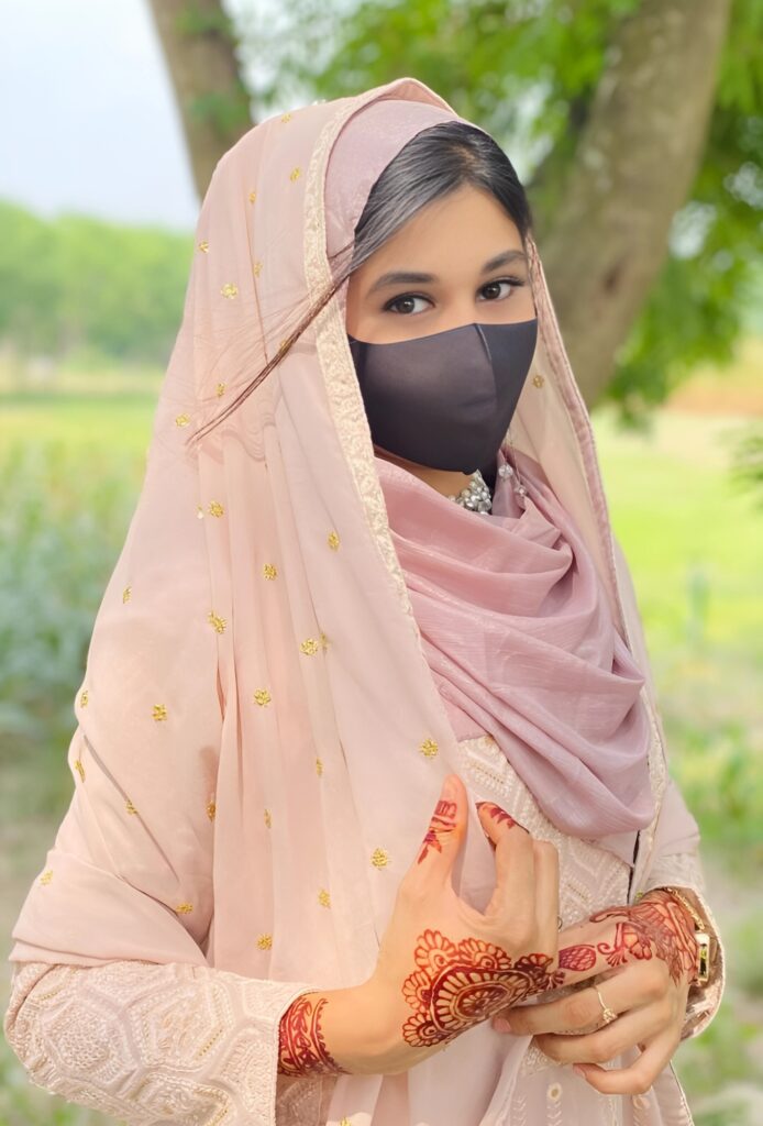 Muslim girl in pastel pink traditional dress with henna-decorated hands and black face mask, standing outdoors near trees