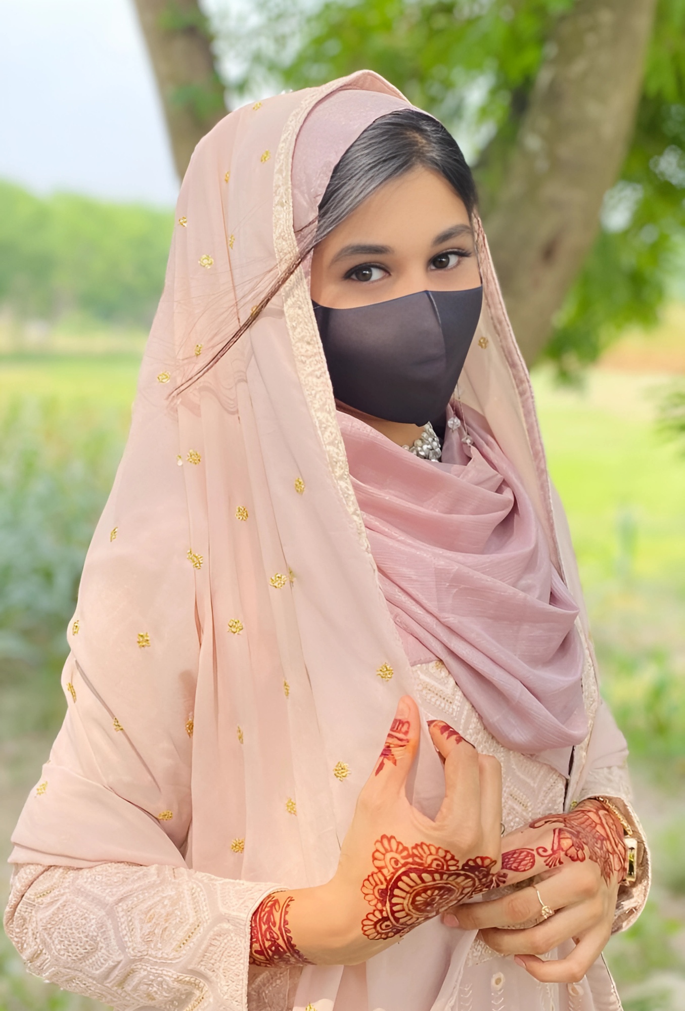 Muslim girl in pastel pink traditional dress with henna-decorated hands and black face mask, standing outdoors near trees