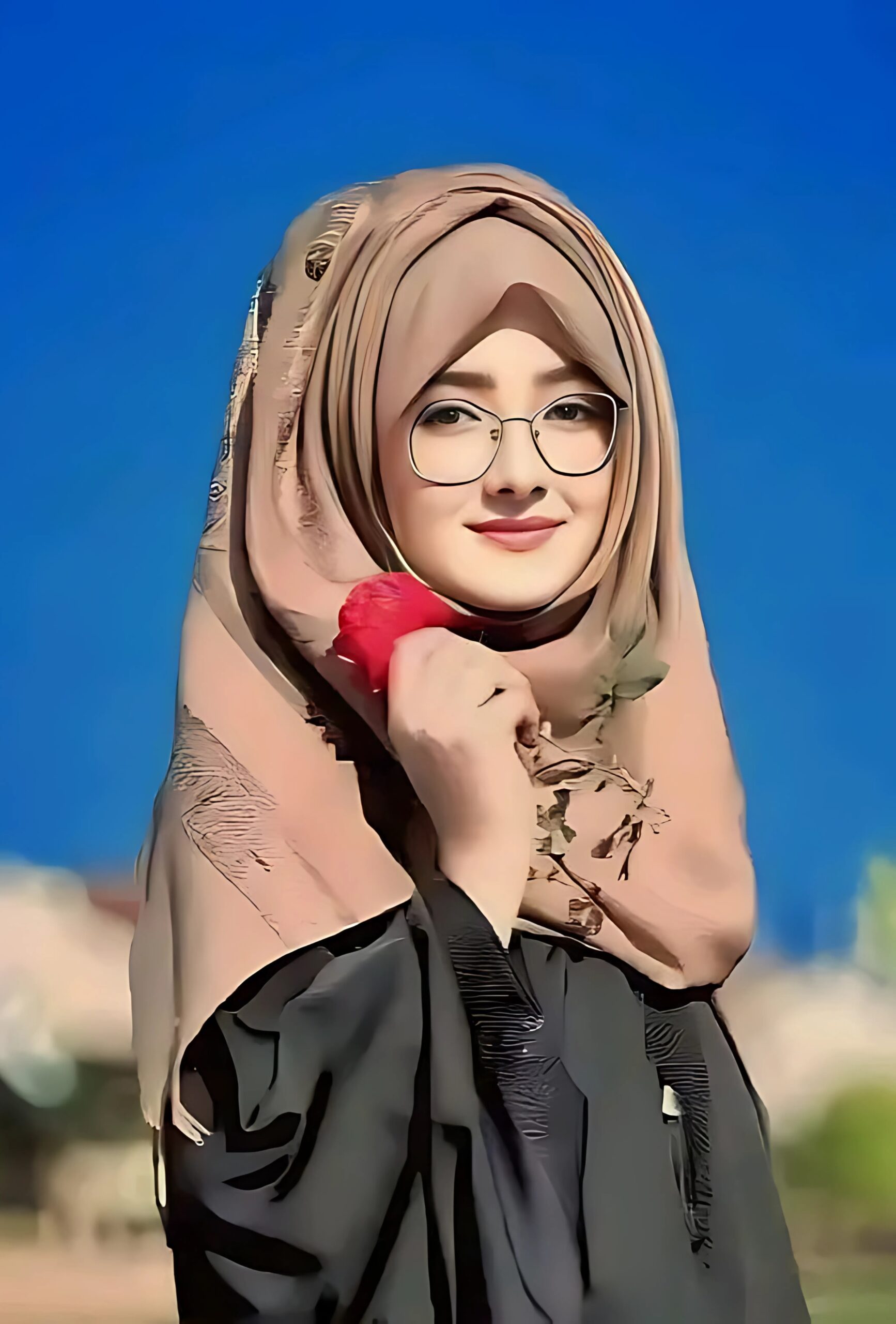 Smiling Muslim girl in brown hijab and glasses holding a red rose against a clear blue sky