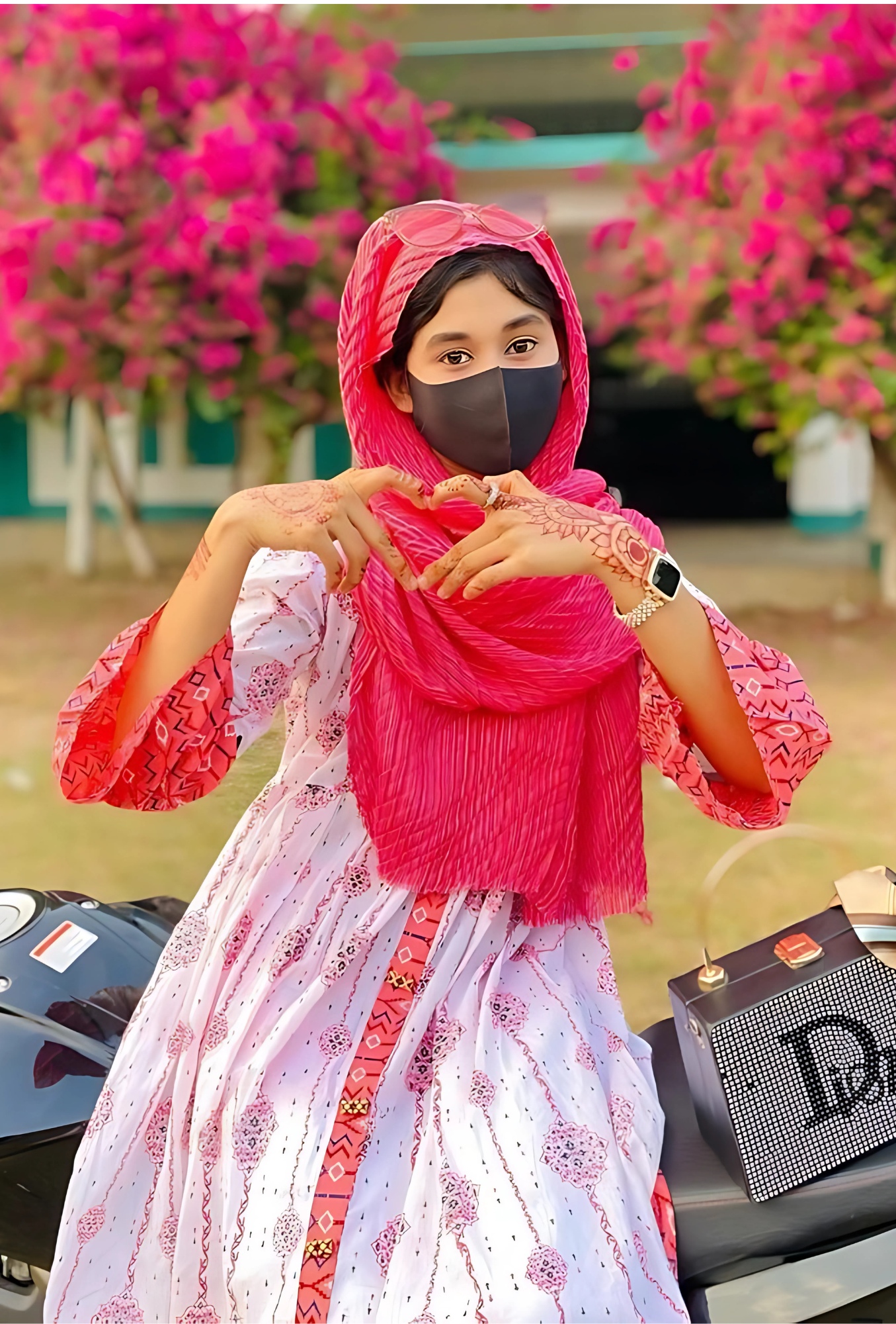 Muslim girl in white and red traditional outfit with black face mask and henna on hands, sitting on a bike with blooming pink flowers in the background.