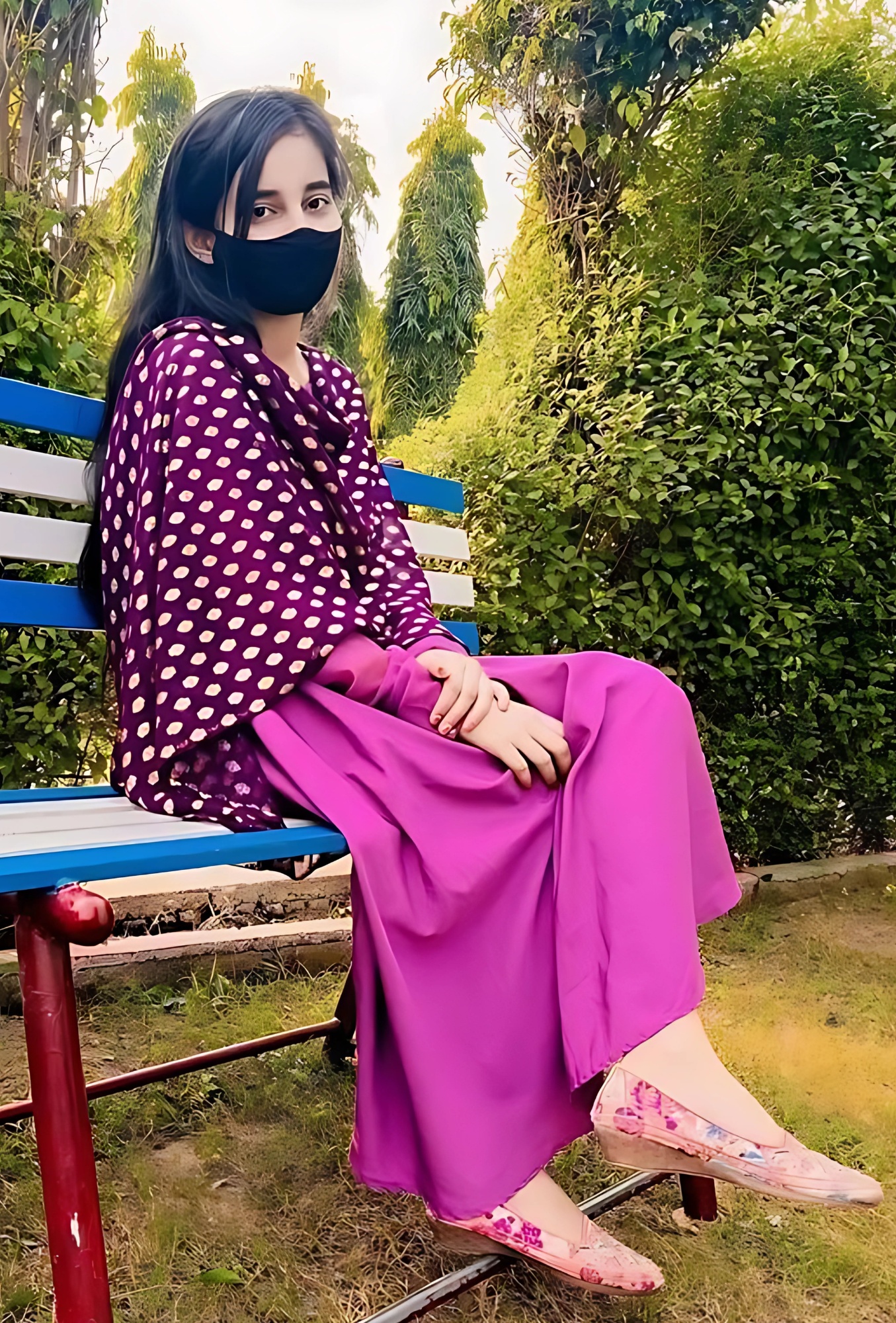 Muslim girl wearing a purple traditional outfit and face mask, sitting on a park bench surrounded by greenery