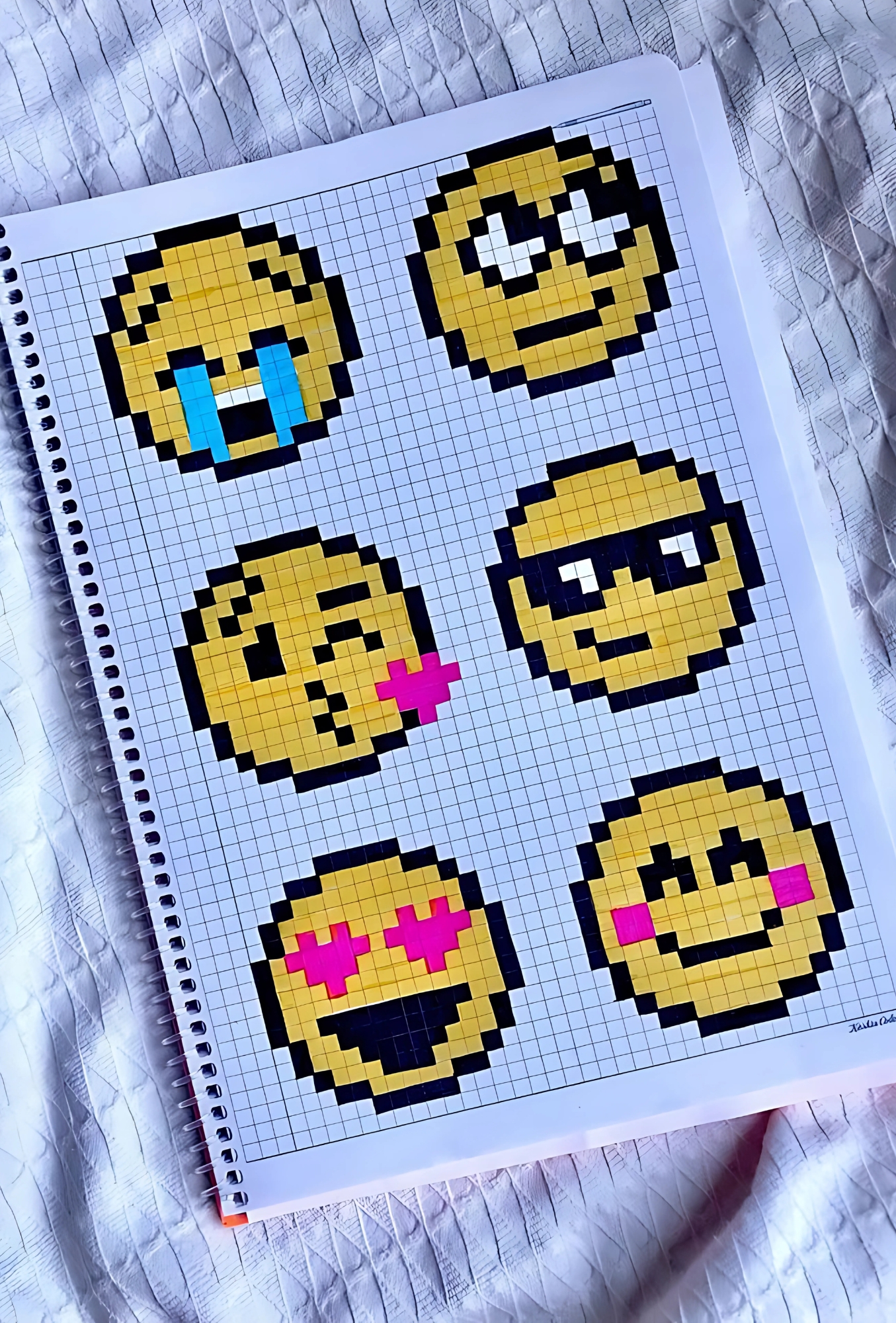 Cuaderno con dibujos pixelados de emojis pequeños y coloridos, incluyendo caritas llorando, enamoradas, con gafas y guiños, hechos sobre papel cuadriculado.