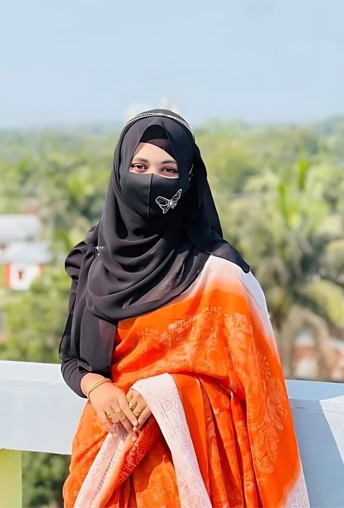 Stylish hijabi girl wearing a black hijab and butterfly mask with an orange traditional saree, posing confidently on a rooftop with a scenic green village backdrop – ideal hijabi girl photo DP.