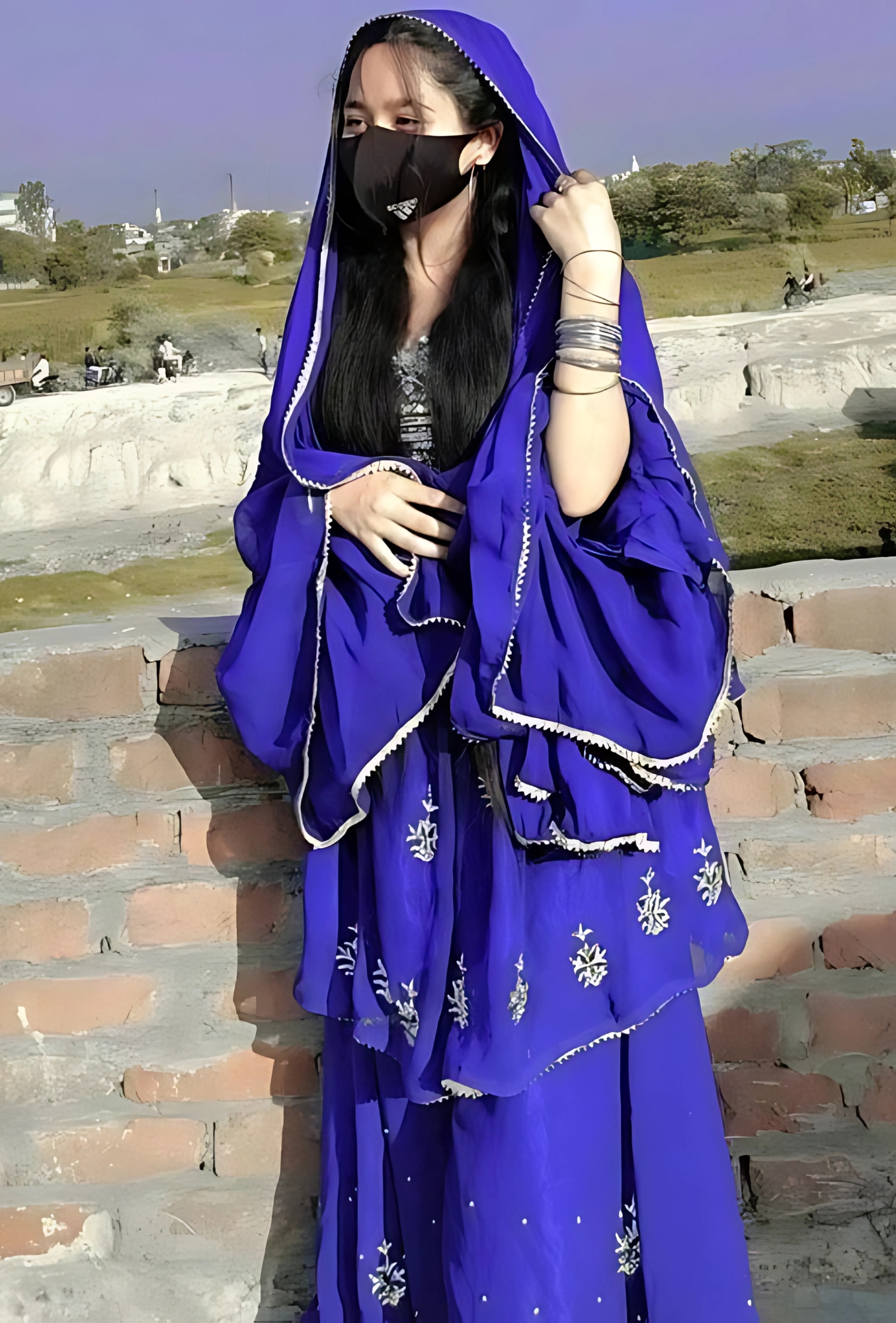 Muslim girl in a royal blue traditional dress wearing a face mask and bangles, standing by a brick wall outdoors